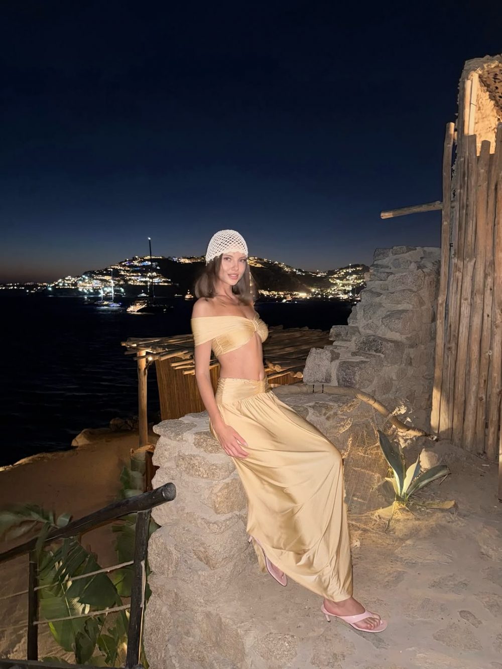 Woman in a beige off-shoulder top and long skirt sits on a stone ledge by the sea at night, with a lit coastal town in the background and a white knit hat.