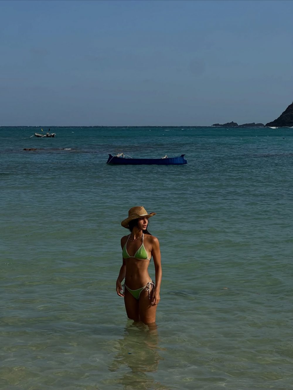 Woman in a lime-green bikini standing in shallow turquoise water, wearing a straw hat, with a boat and distant rocky shore in the background