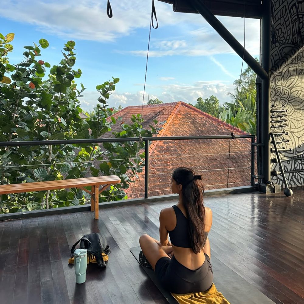 Woman in a black sports top and shorts sits cross‑legged on a yoga mat on a wooden balcony, facing greenery and a red-tiled roof.