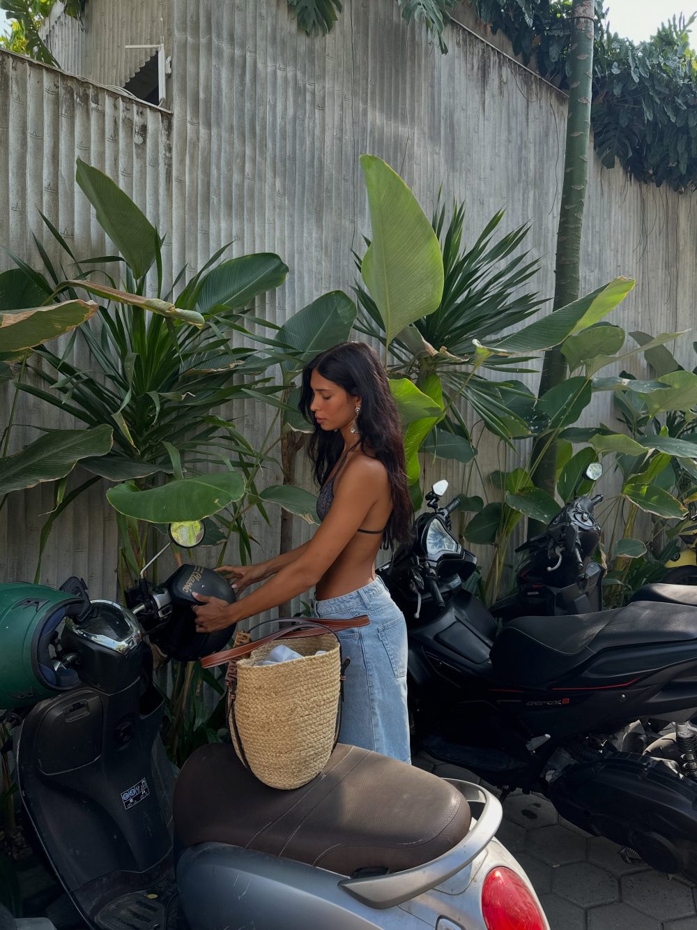 Woman with long dark hair in a gray top and jeans stands beside parked motorbikes, holding a helmet, near tropical plants and a gray wall.