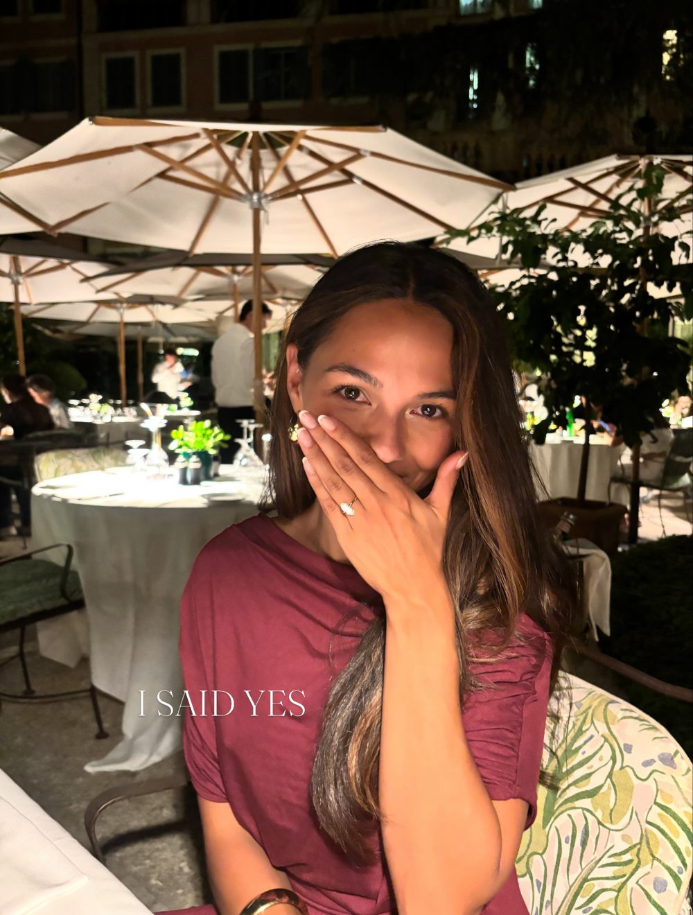 Woman in a maroon dress covers her mouth with her hands at an outdoor night-time restaurant; 'I SAID YES' text appears at lower edge.