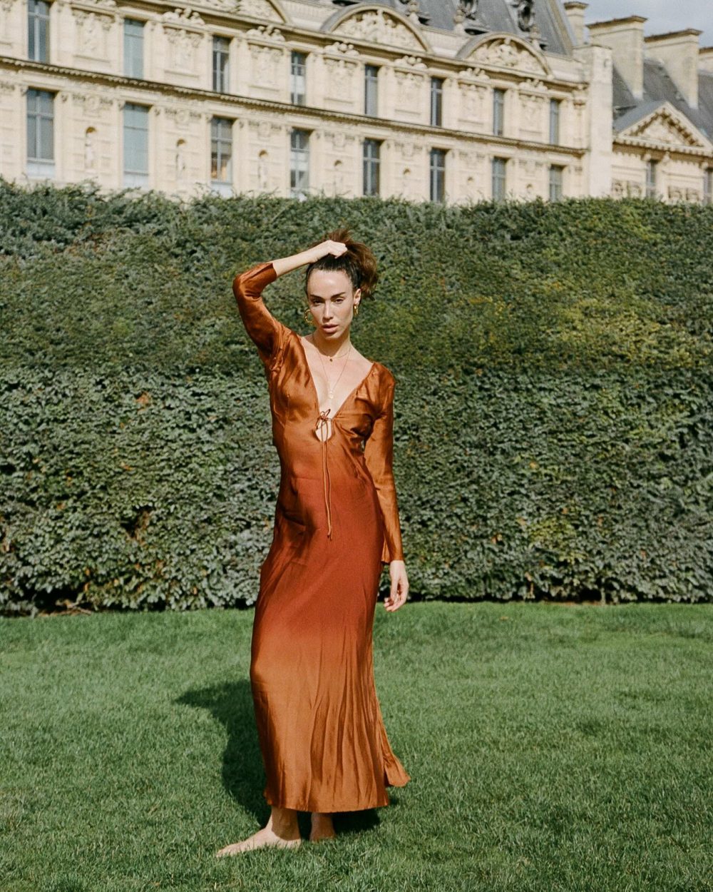 Woman in a rust satin dress standing barefoot on grass, one hand in her hair, with a hedged backdrop and ornate historic building behind her.
