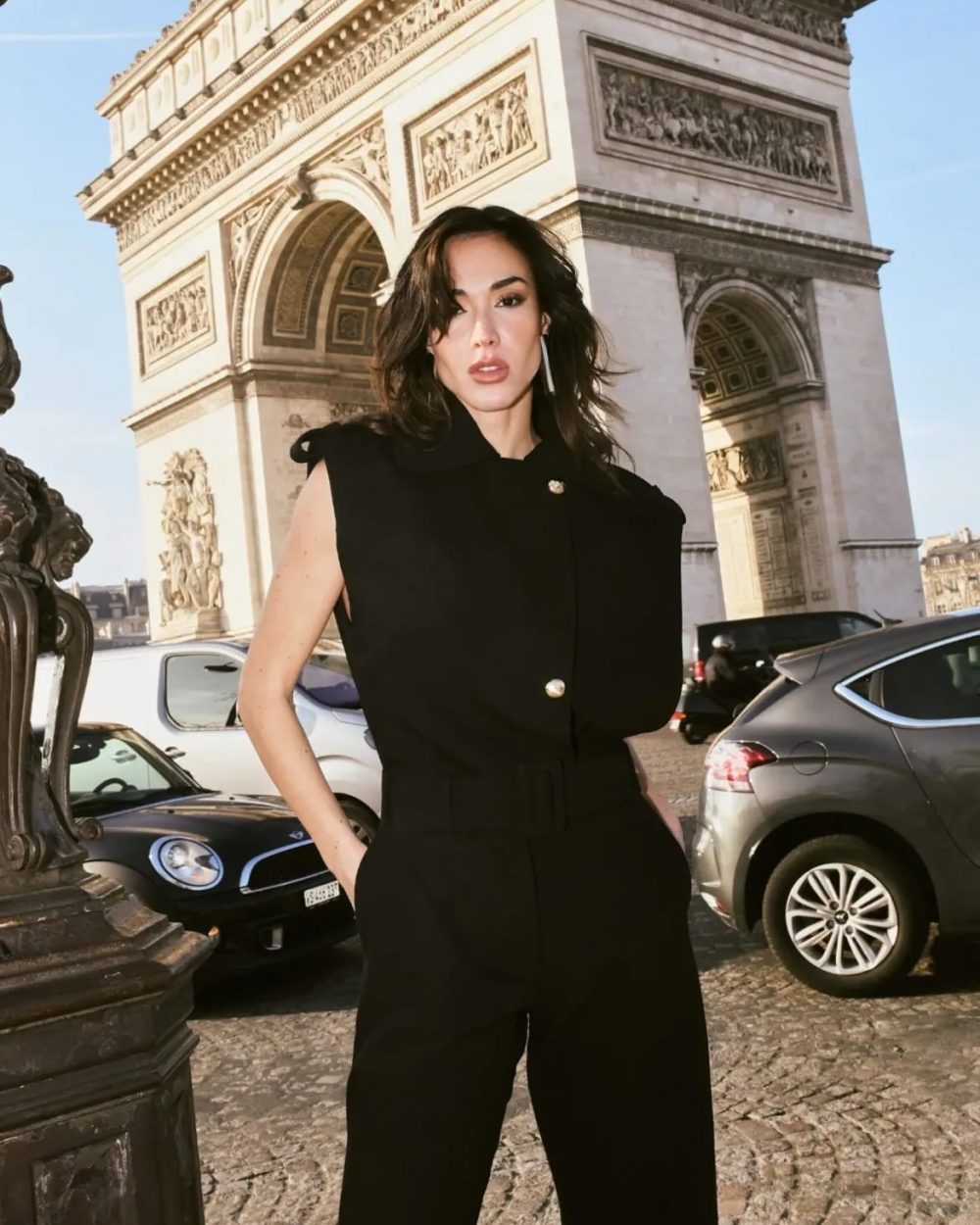 Woman in a black sleeveless jumpsuit standing on a Paris street in front of the Arc de Triomphe du Carrousel, with cars and cobblestones in the background