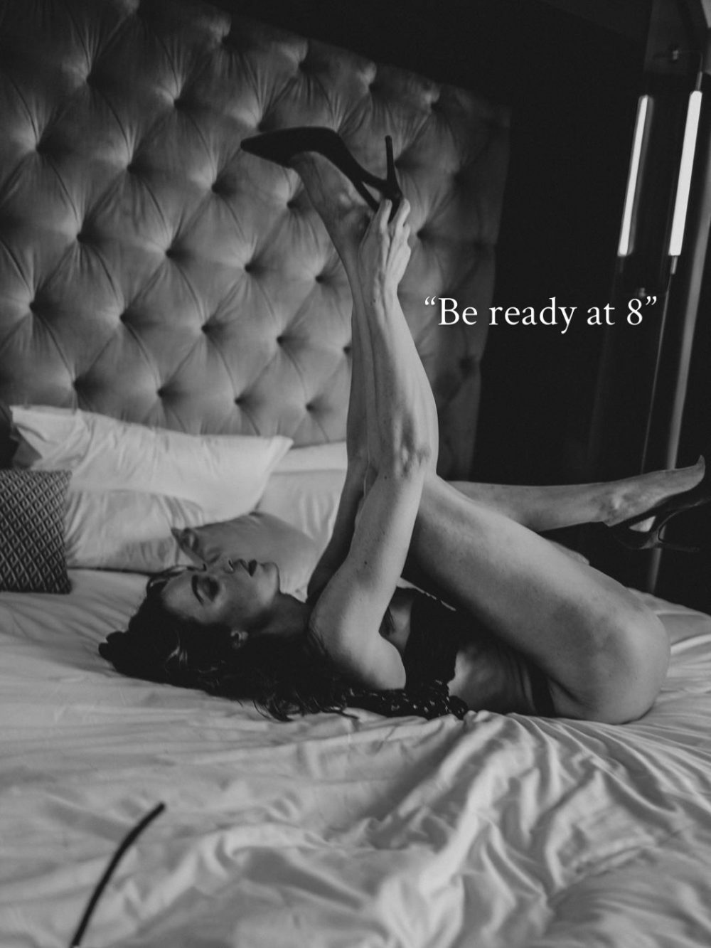 Person lying on a bed with legs raised in the air wearing high heels; a caption reads “Be ready at 8.”