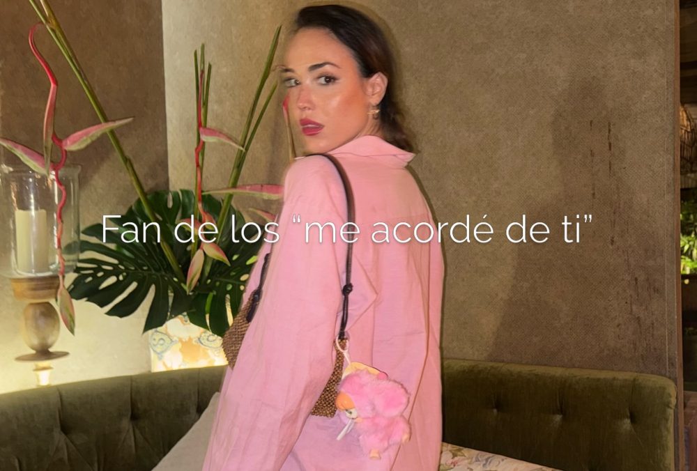 Mujer de espaldas con abrigo rosa claro en un salón, rodeada de plantas y una vela decorativa; mirada hacia la cámara.