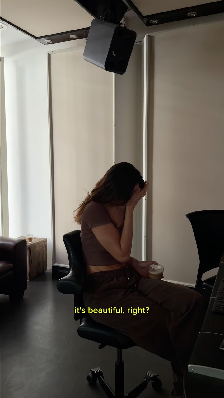 Woman seated in an office chair in a studio, one hand covering her face, holding a cup with a laptop nearby.