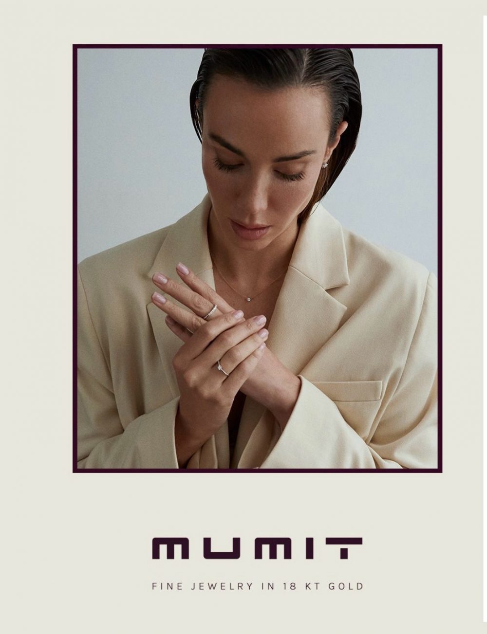 Portrait of a woman in a beige blazer looking down, hands held together showing multiple rings; jewellery ad for Mumit 18K gold