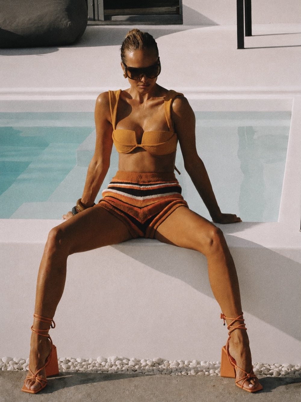 Woman in an orange bikini top and striped orange shorts sits poolside on a white ledge, wearing sunglasses.