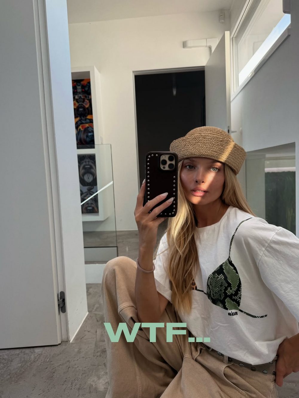 Woman taking a mirror selfie in a bright, minimalist room, wearing a straw hat, white graphic tee, and beige pants, with a 'WTF..' overlay text.