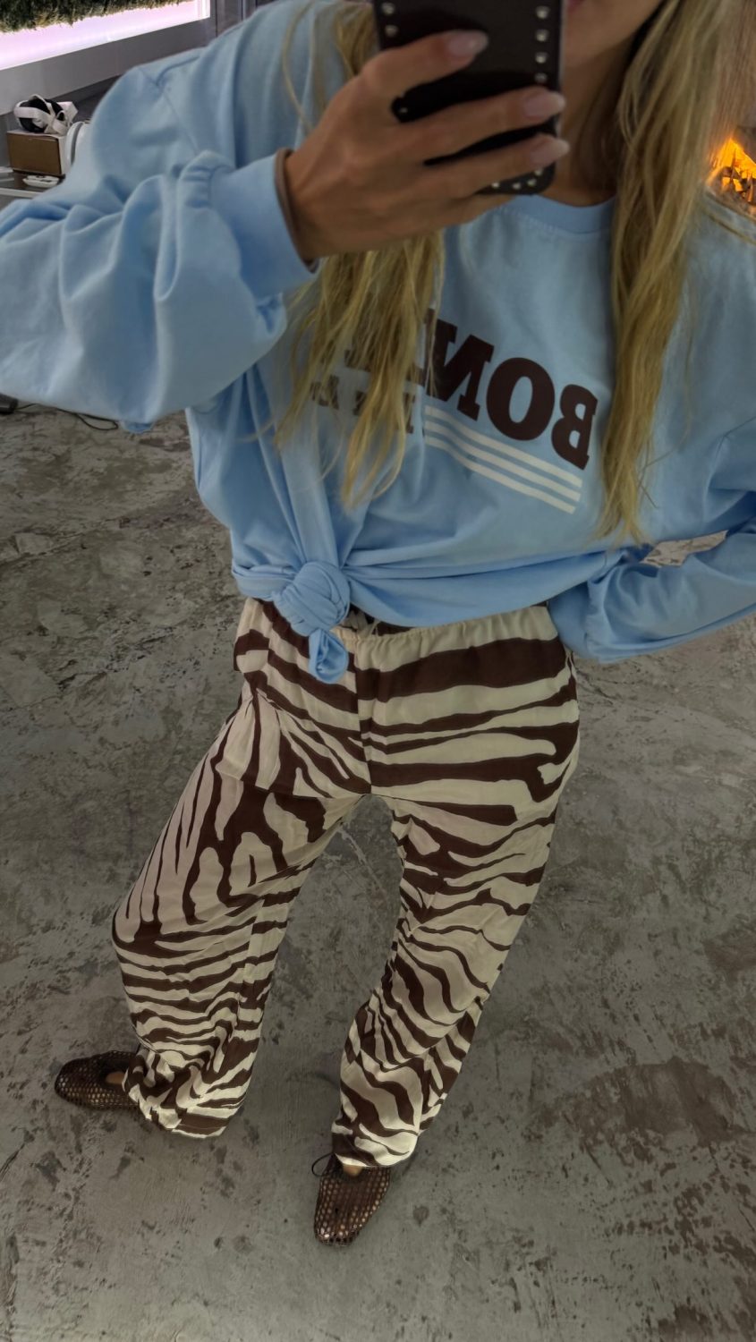 Person taking a mirror selfie wearing a light blue tied sweatshirt and bold zebra-print pants, with brown woven shoes indoors near a fireplace.