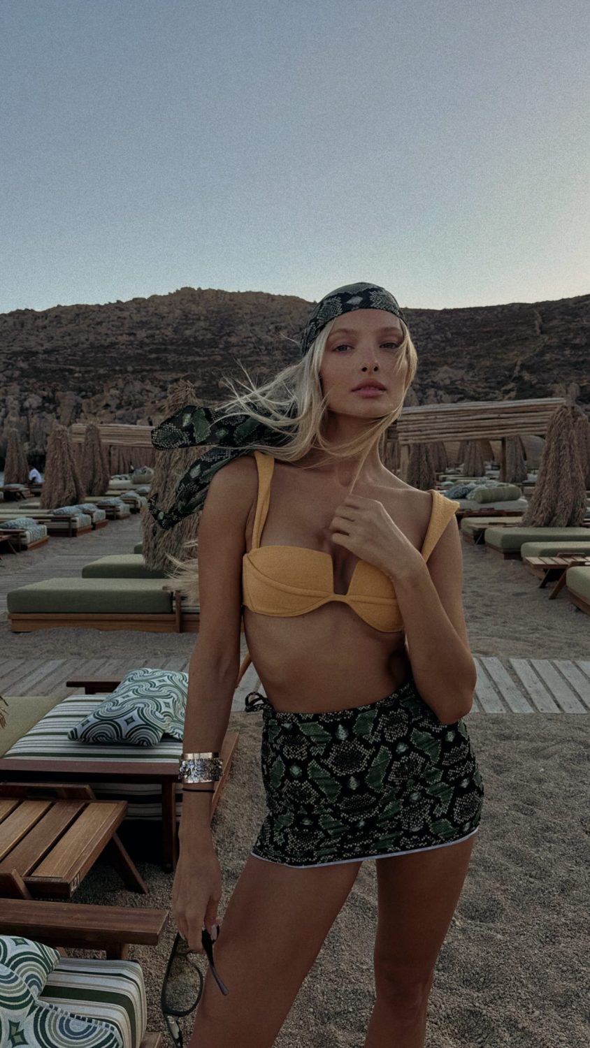 Young woman at a beach club wearing a yellow bikini top and green snake-print skirt, wind blowing her hair as she holds sunglasses nearby.