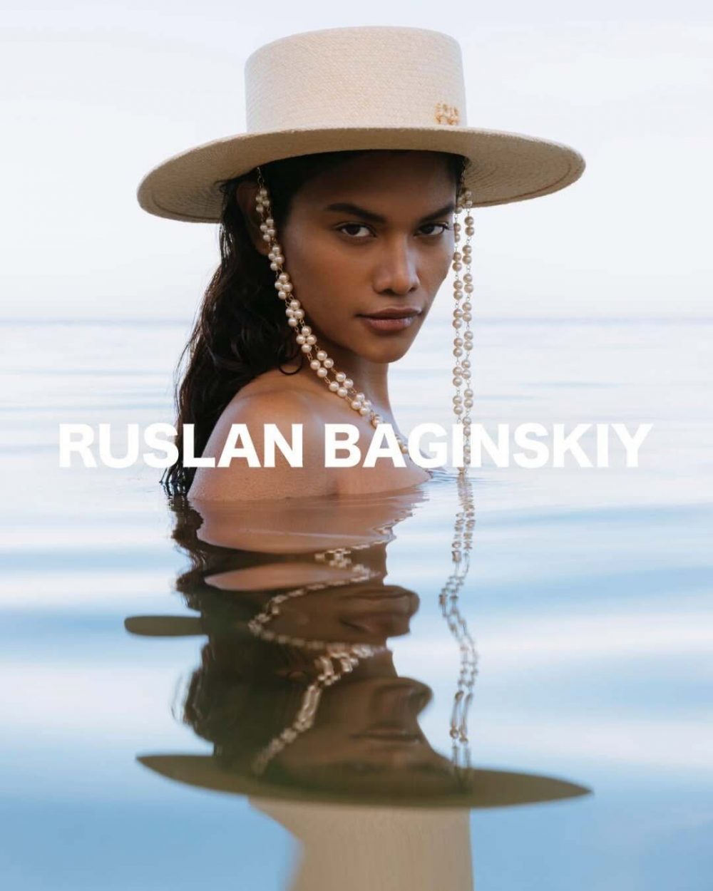 Woman in a wide-brim straw hat and pearl necklace, half-submerged in water, with the text 'RUSLAN BAGINSKIY' across.