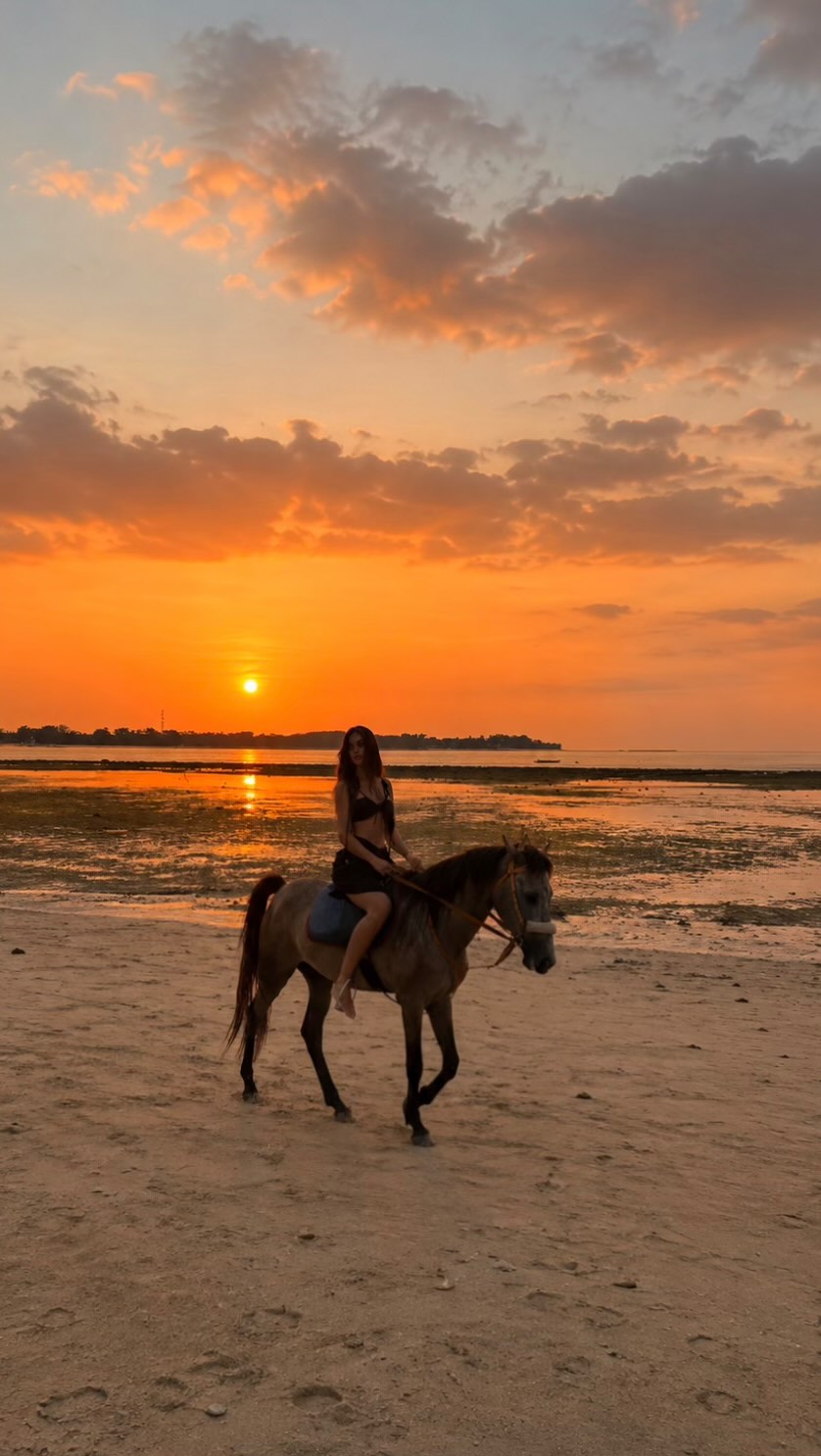 Woman riding a horse along a sandy beach at sunset, orange sky and calm water in the background.