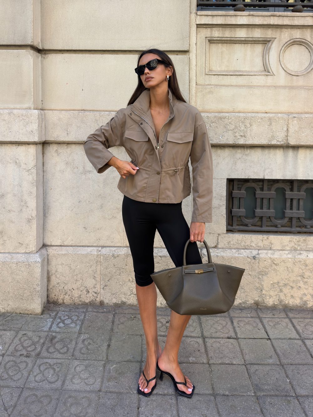 Stylish woman in a beige utility jacket, black leggings, and flip-flop sandals stands against a stone wall, holding a large taupe handbag.