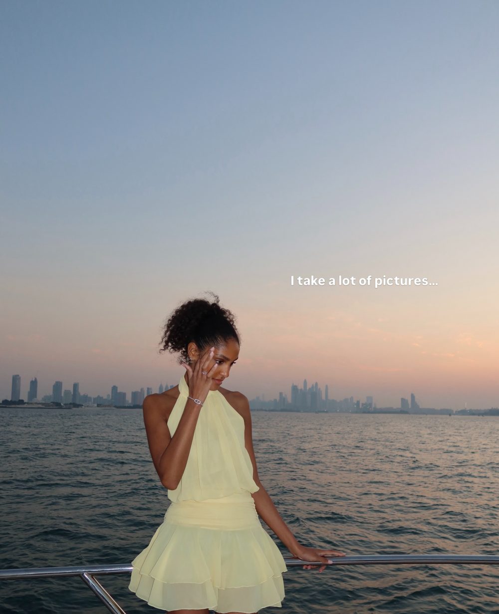 Woman in a pale yellow dress standing at a boat railing over the water, with a city skyline at sunset in the background (caption: 'I take a lot of pictures...').