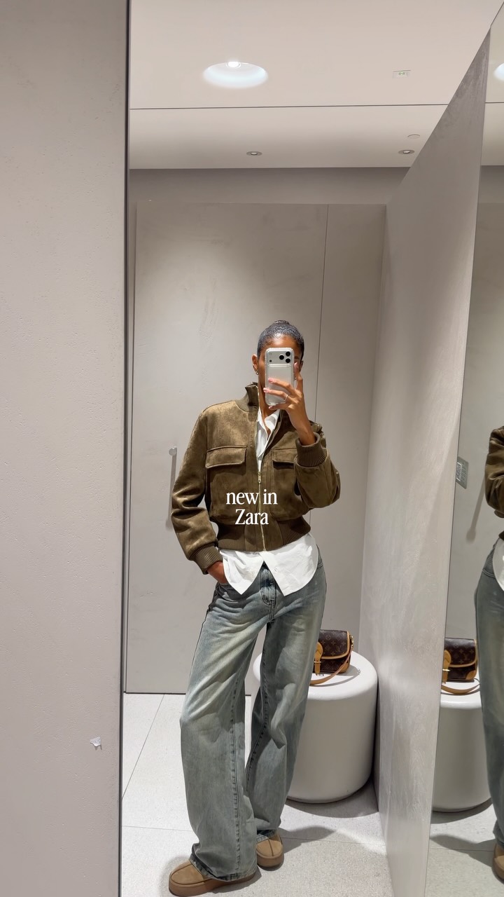 Person taking a full-length mirror selfie in a clothing store, wearing a brown bomber jacket over a white shirt and light-wash jeans; a brown purse sits on a white round stool nearby.