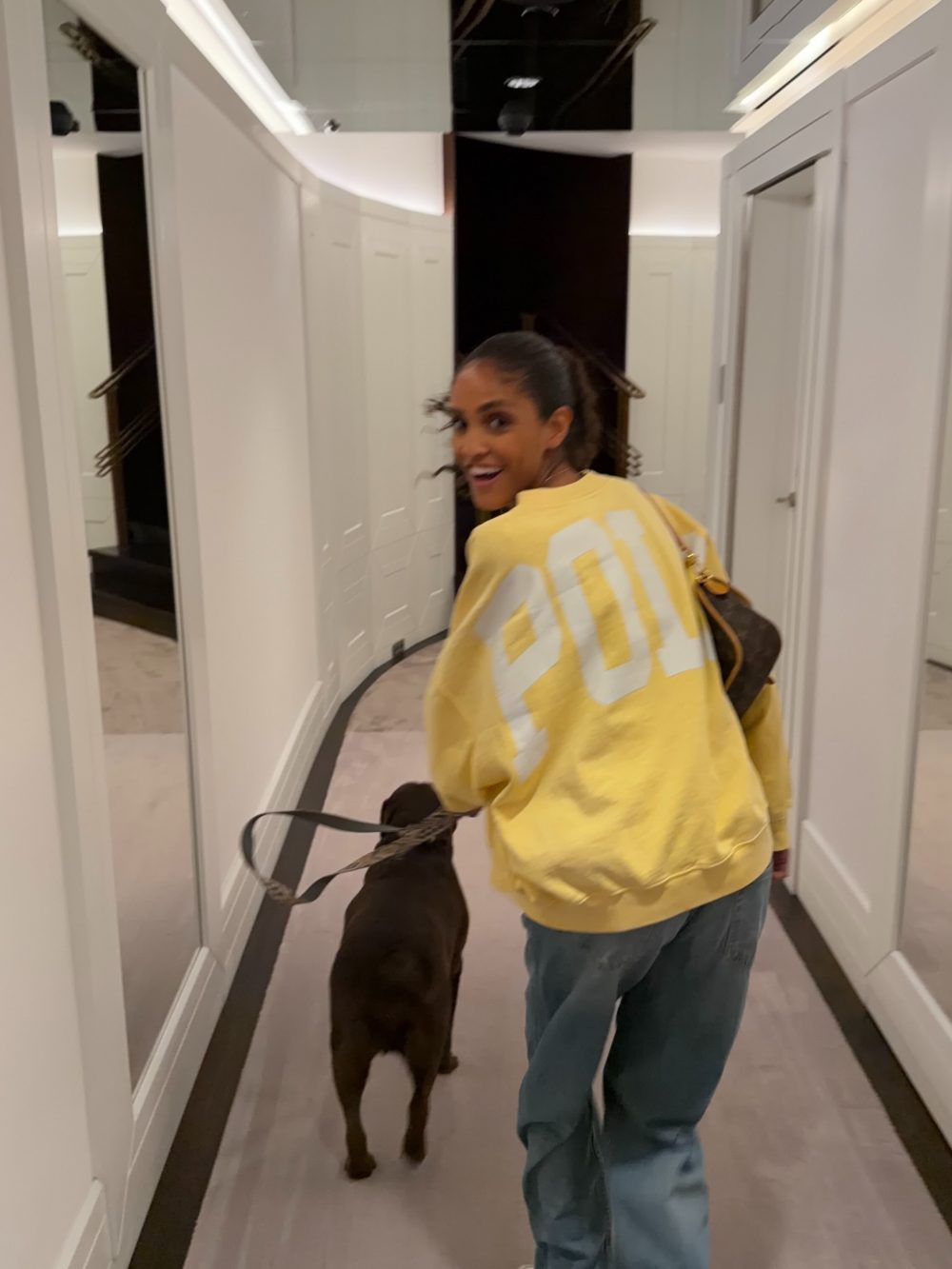 Woman wearing a yellow sweatshirt with large white numbers on the back, smiling and walking a brown dog on a leash down a white hallway with lockers on the left.