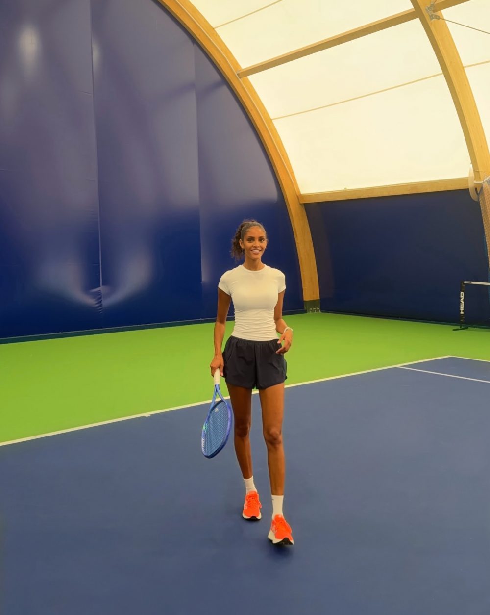 Female tennis player standing on blue-green indoor court holding a blue racket, wearing a white shirt, black shorts, and bright orange shoes.