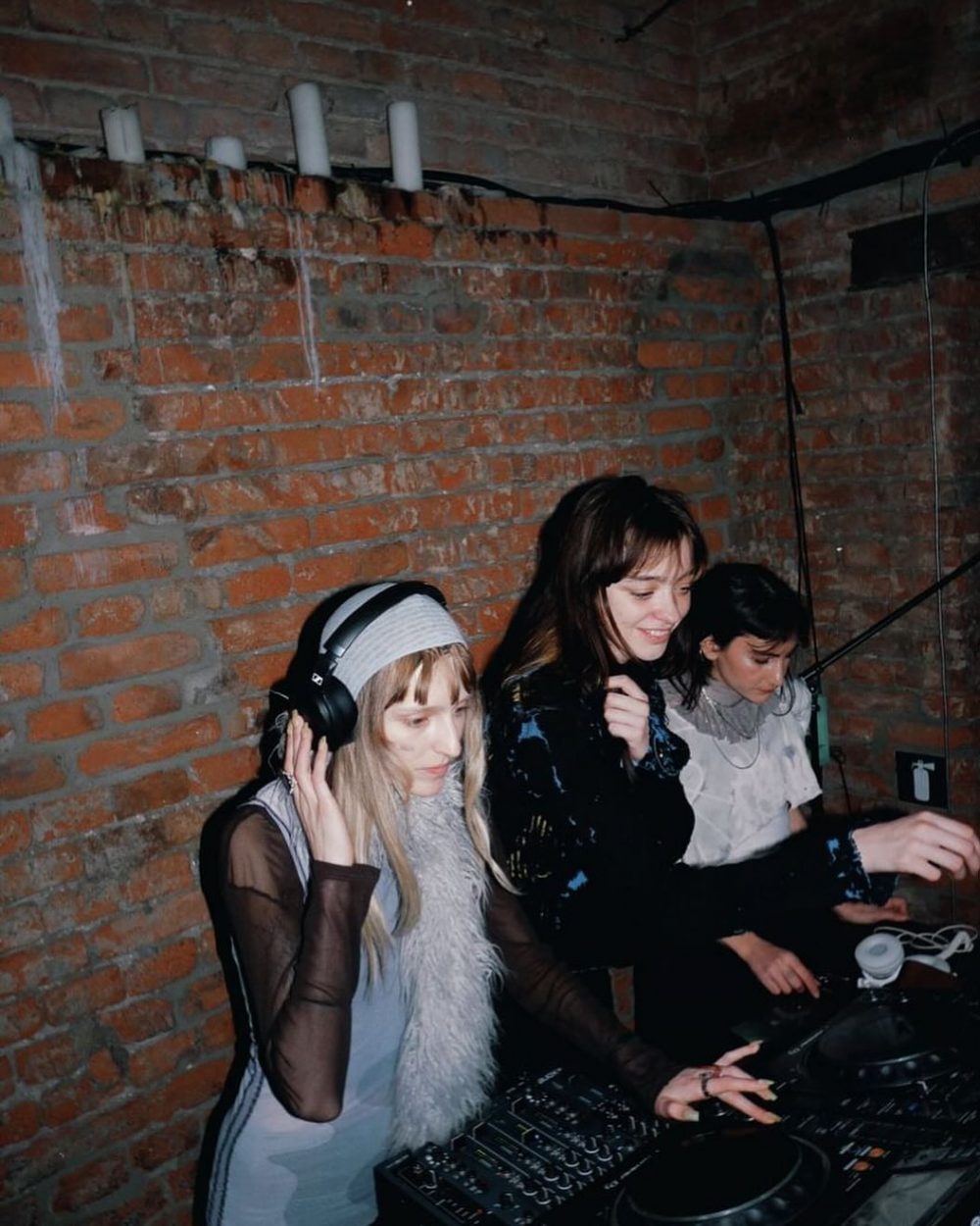 Three women earnestly DJ in a brick-walled basement, wearing headphones and mixing on a DJ mixer and turntables.