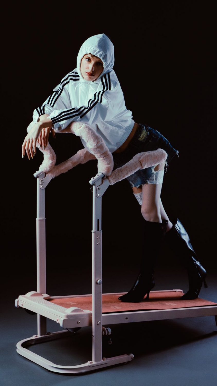Person in a white hooded jacket and ripped denim shorts leans on a pink-furred, treadmill-style exercise machine in a dark studio background.