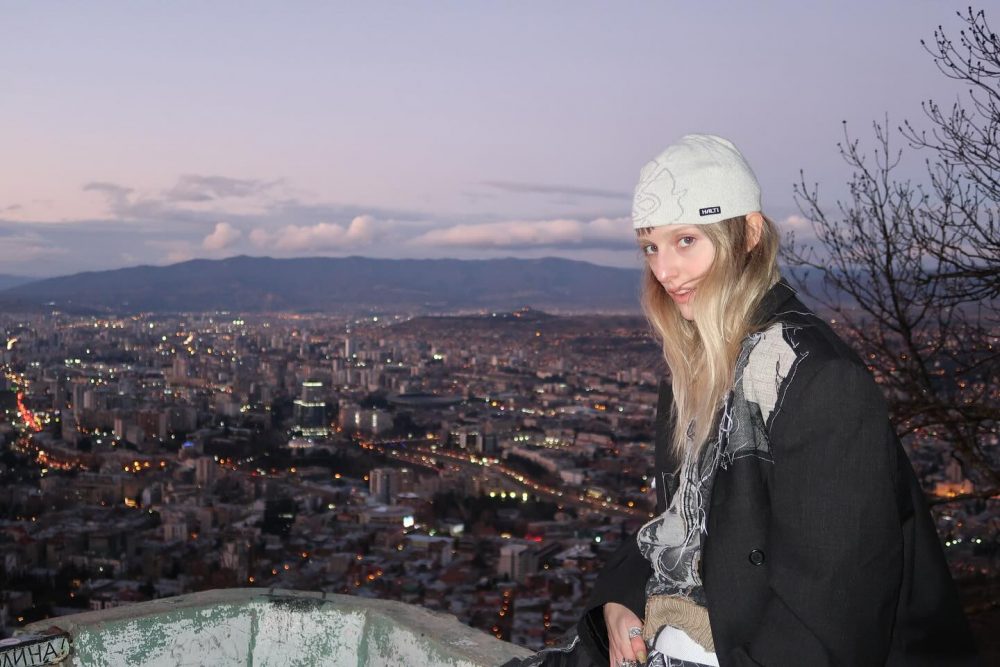 Woman in a light beanie and black coat sits on a hill, overlooking a lit city at dusk with mountains in the distance.