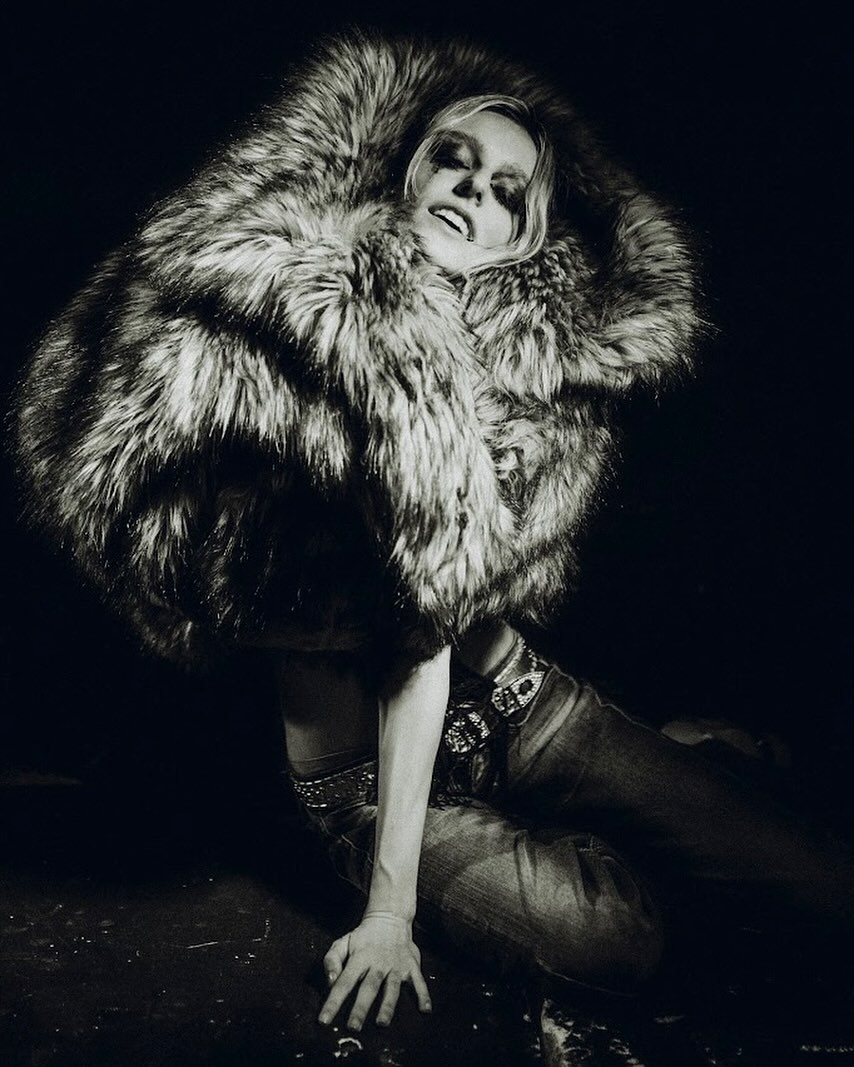 Person in a voluminous fur coat crouches on the ground, smiling with eyes closed in a dramatic fashion shot.
