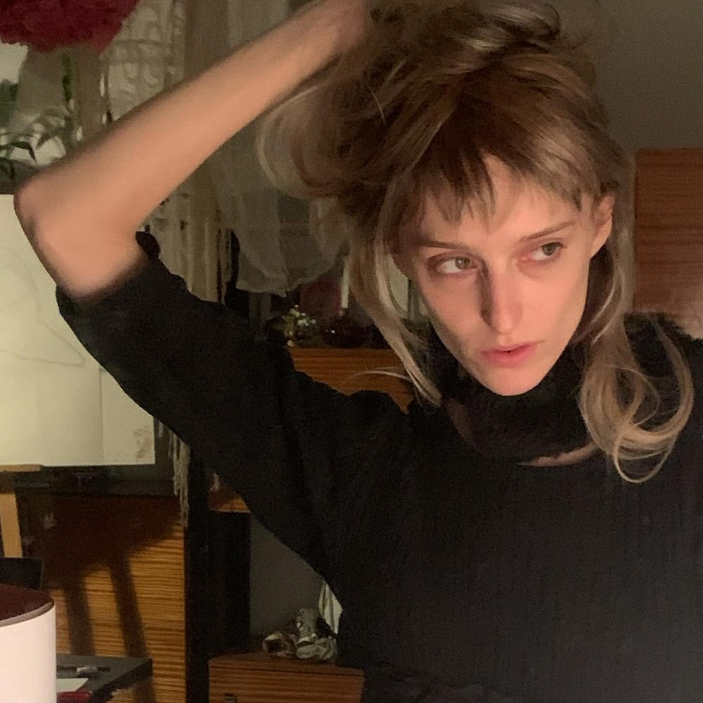 Person with brown-blonde hair in a black top lifting hair with one hand in a dimly lit room, looking off to the side.