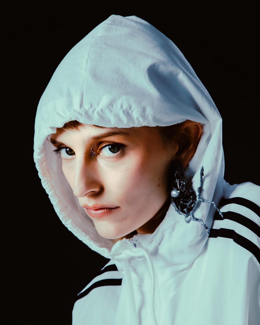 Portrait of a person in a white hooded jacket against a black background, with a metallic earring visible on the left ear.