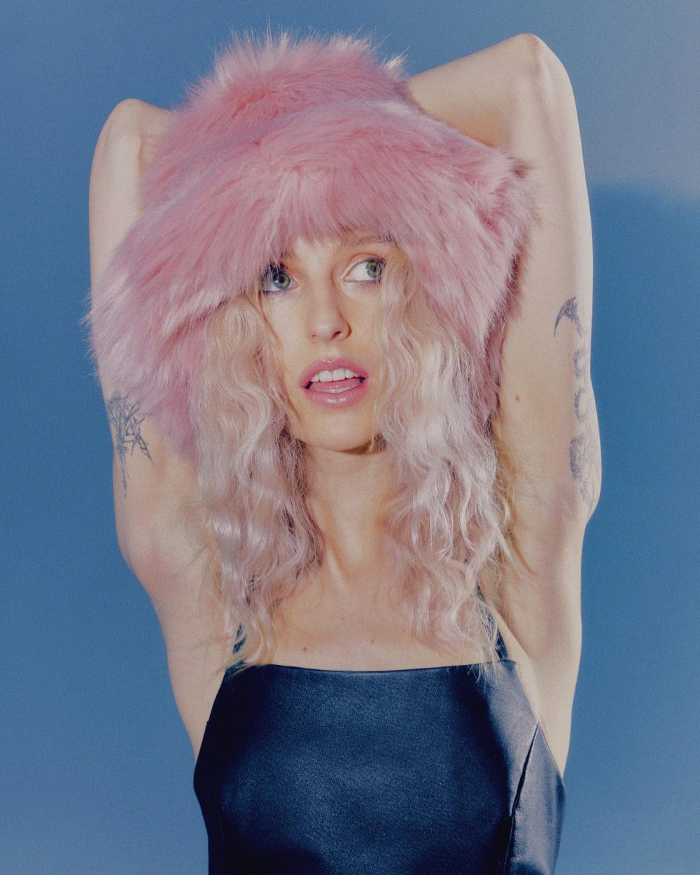 Woman with pastel pink hair, wearing a dark satin top, arms raised behind her head against a blue studio backdrop, tattoos visible on arms.