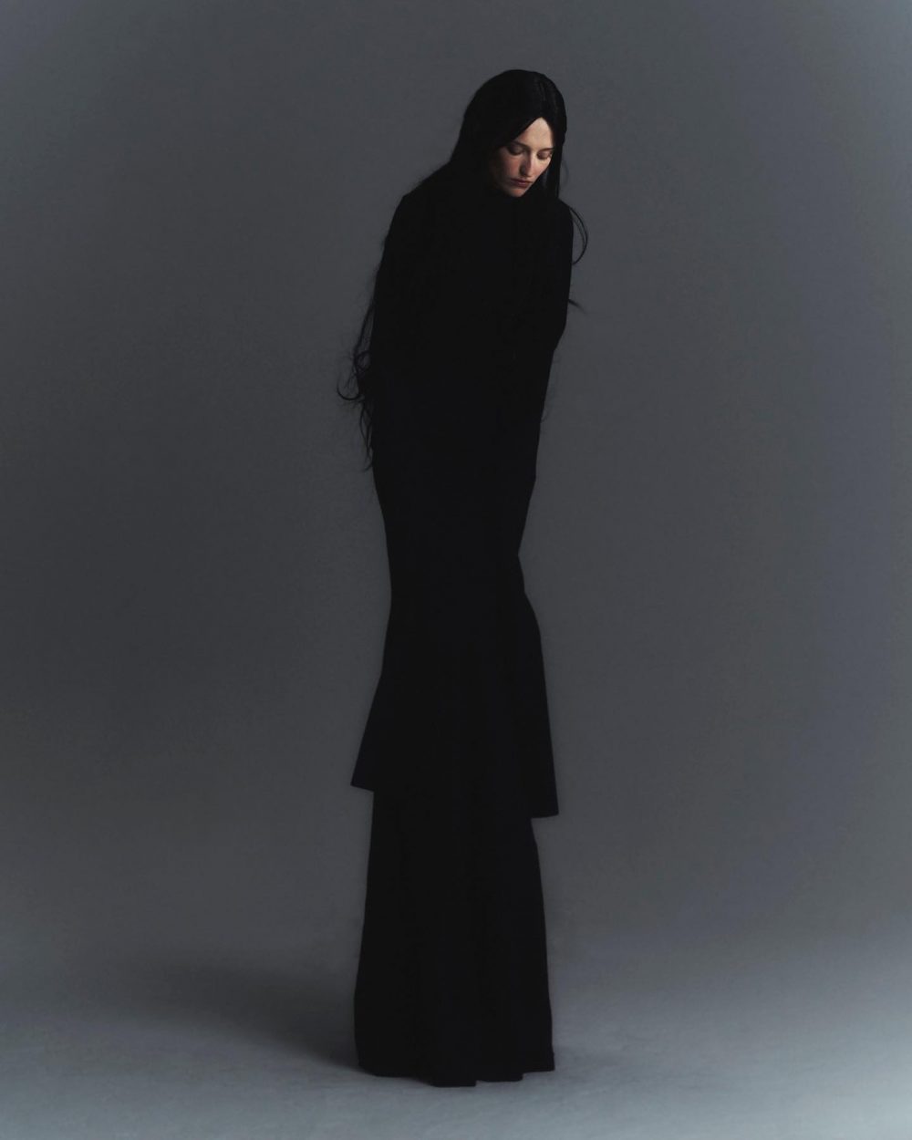 Woman wearing a long black dress standing against a gray backdrop, looking down with a somber expression.