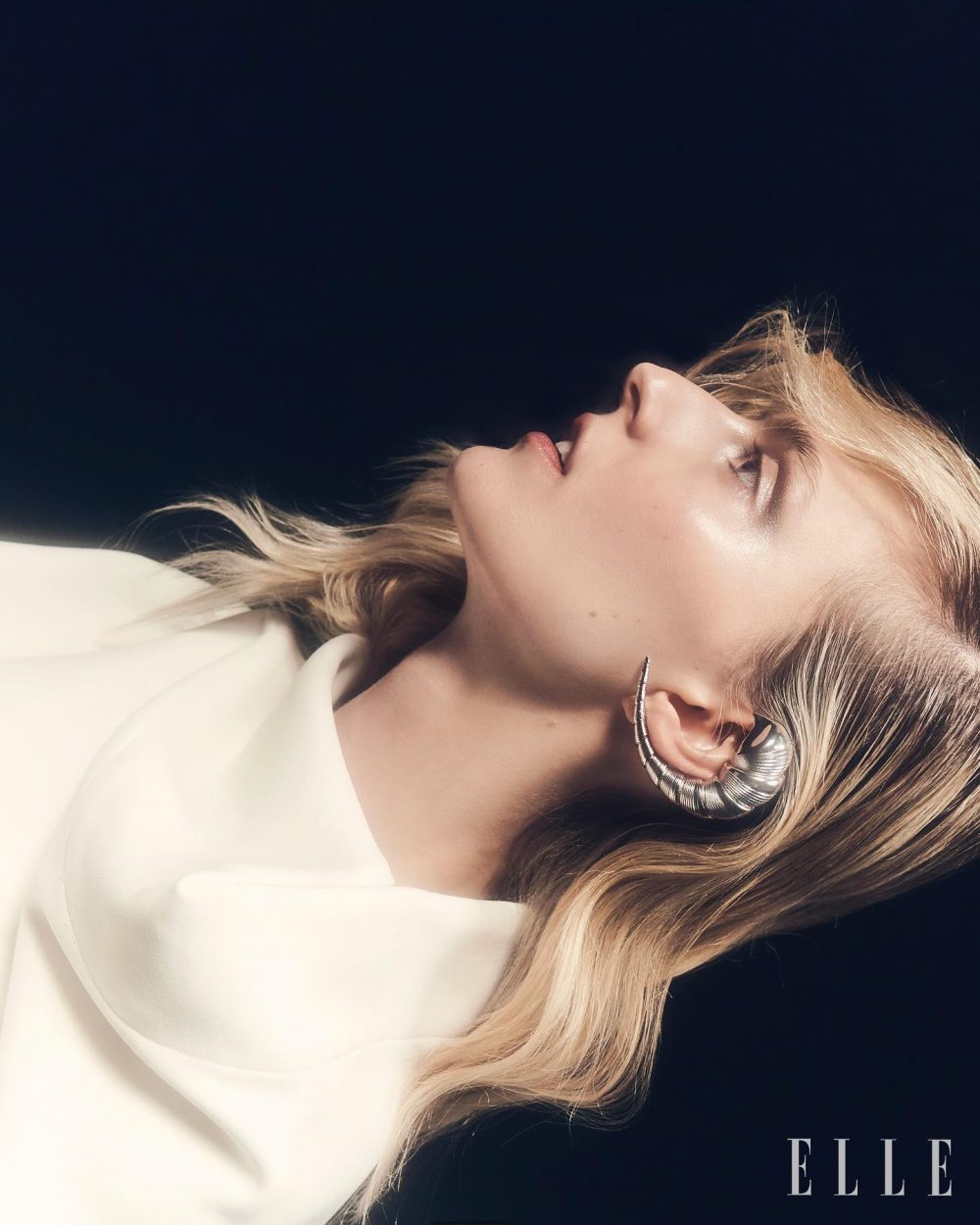 Blonde woman in a white blouse, head tilted back, wearing a thick silver hoop earring against a dark background.