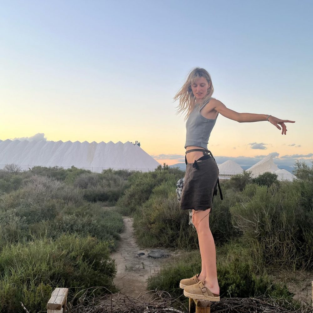 Woman balancing on small wooden blocks outdoors at sunset, arms extended, with salt miles in the background.