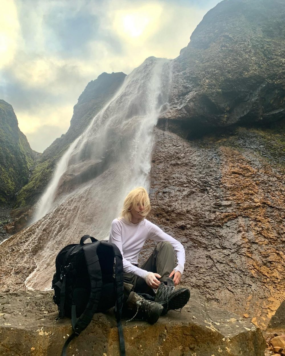Person with blonde hair sits on wet rocks beside a waterfall, wearing a white long-sleeve shirt and green pants, with a black backpack nearby.