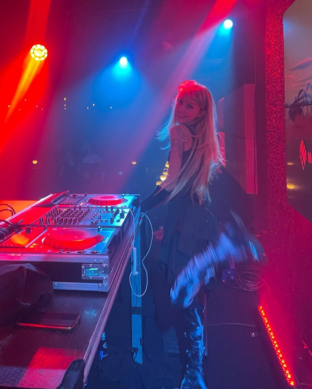 Female DJ with long blonde hair at a mixing desk, colorful red and blue stage lights overhead, nightclub scene behind her no audience visible in focus, musical equipment in foreground.