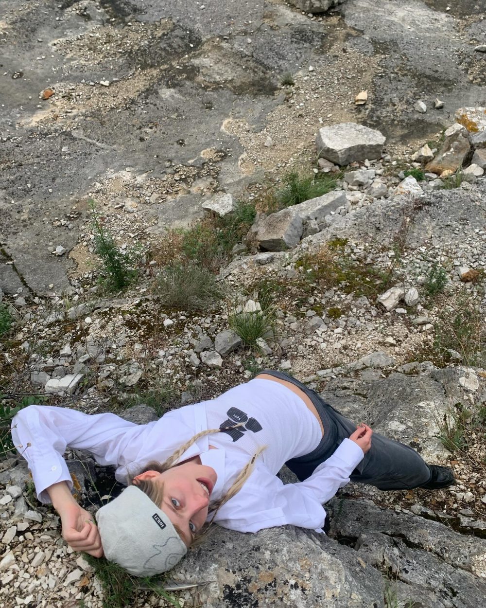 Person lying on a rocky, sparse hillside wearing a white shirt and gray beanie, looking up at the camera.
