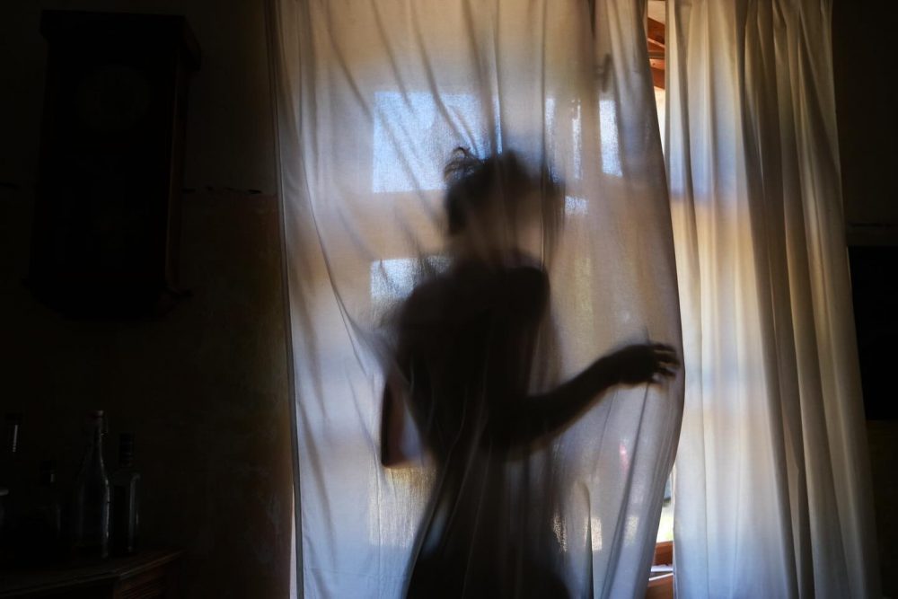 Person silhouette behind a sheer curtain, backlit by daylight in a dim room.