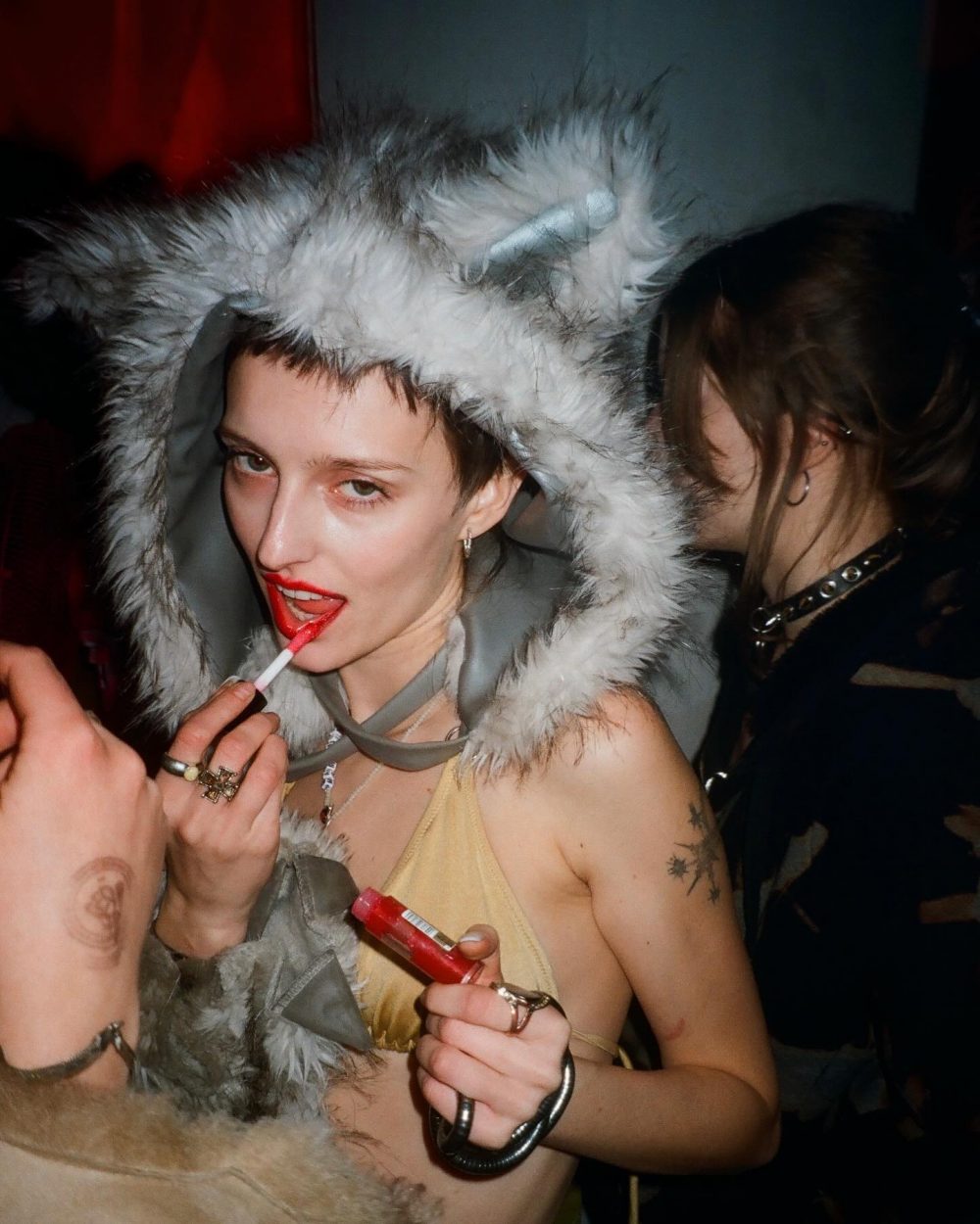 Person wearing a fluffy gray hood applies red lipstick to someone at a party.