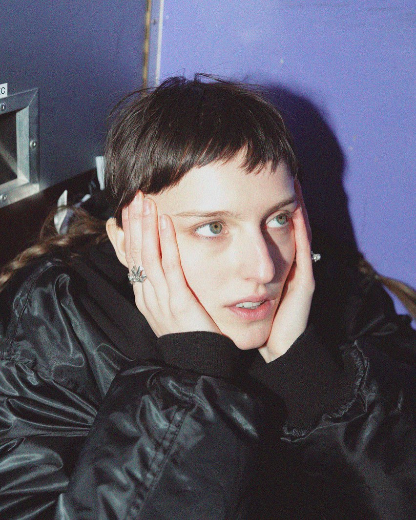 Person with short dark hair rests their face in both hands against a purple wall, looking off to the side.