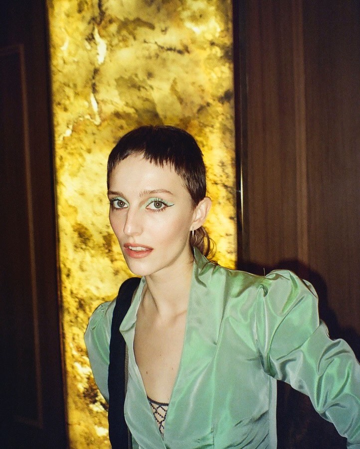 Woman with a short dark pixie cut in a mint-green satin blazer, posing in front of a golden textured wall in a dim interior.