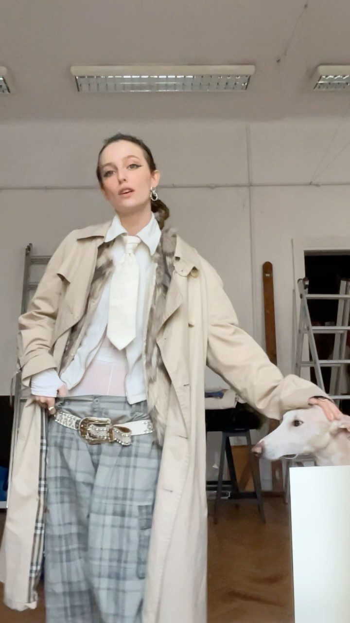 Woman in a beige trench coat and white shirt with a tie, posing in a studio beside a light-colored greyhound.