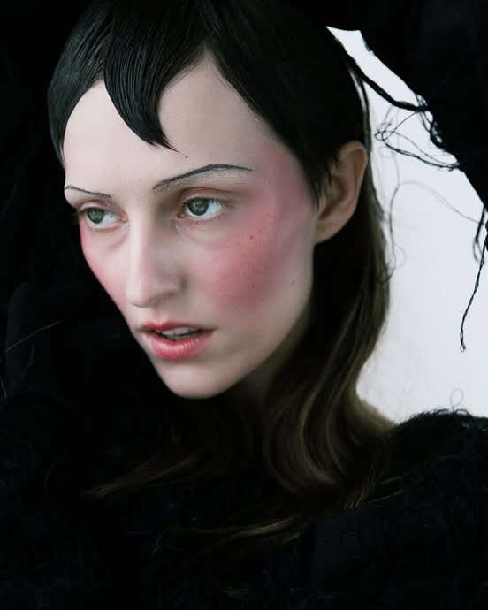 Close-up of a pale mannequin head with black hair and prominent rosy cheeks, set against a dark garment.