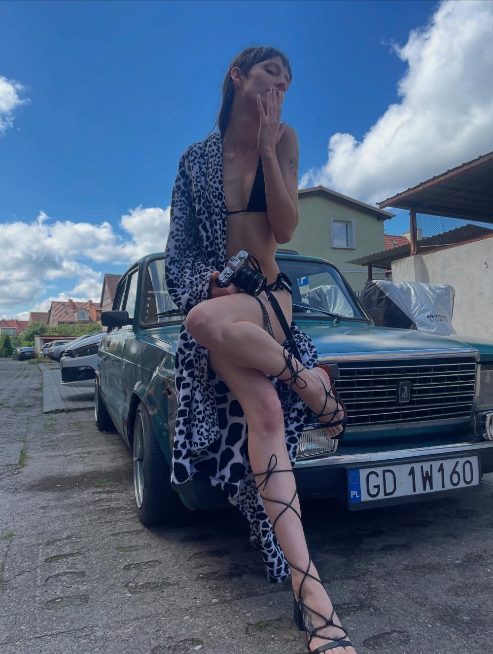 Woman in a black bikini and leopard robe poses on the hood of a vintage blue car in a sunny street. she wears strappy heels and holds a camera while one leg rests on the bumper, blue sky above.