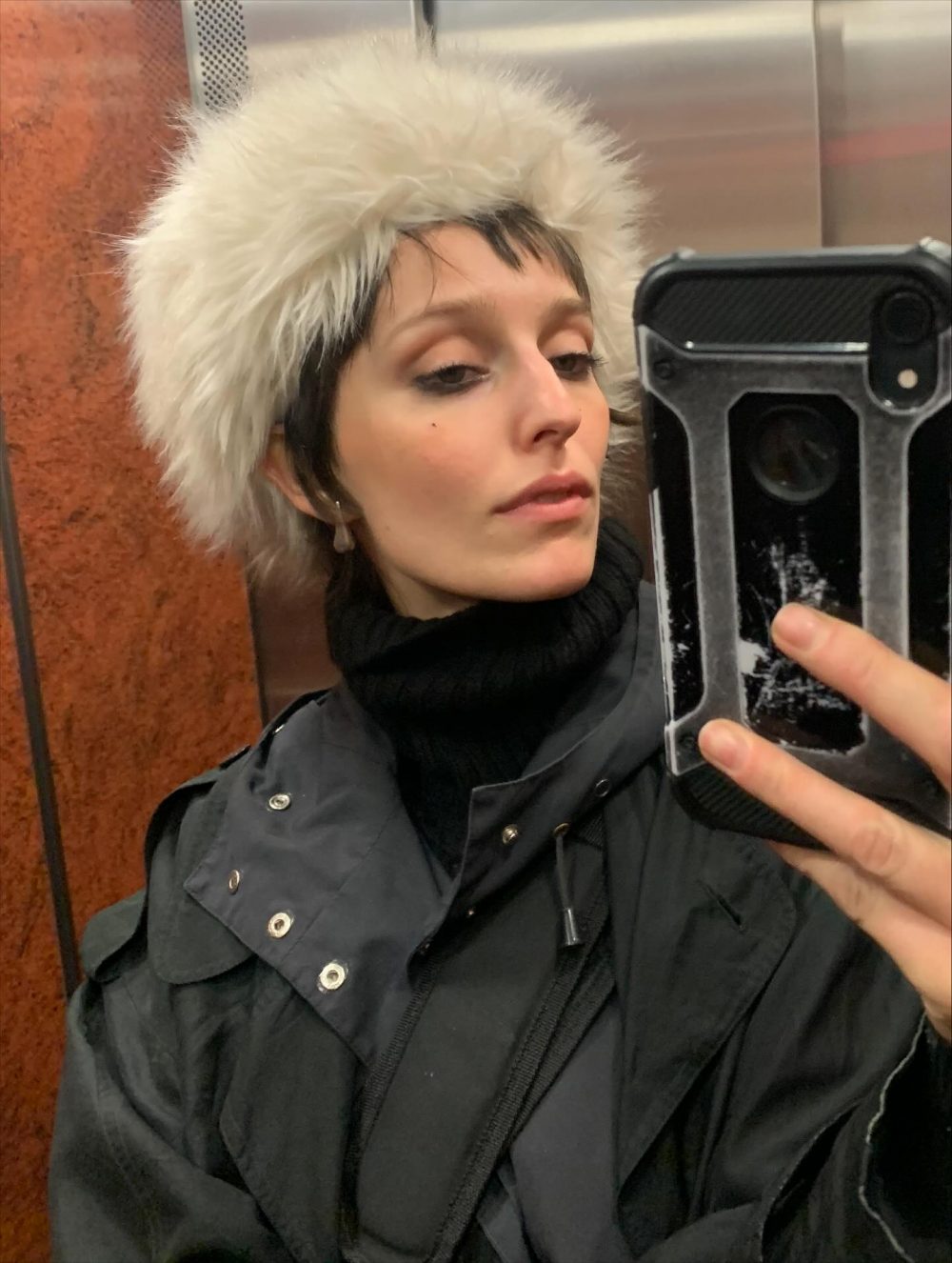 Person taking a mirror selfie in an elevator, wearing a fluffy white fur hat and a black jacket.