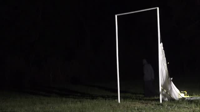 Nighttime outdoor scene: a white rectangular frame standing in a grassy field with a white cloth draped on one side, creating a ghostly silhouette in darkness.