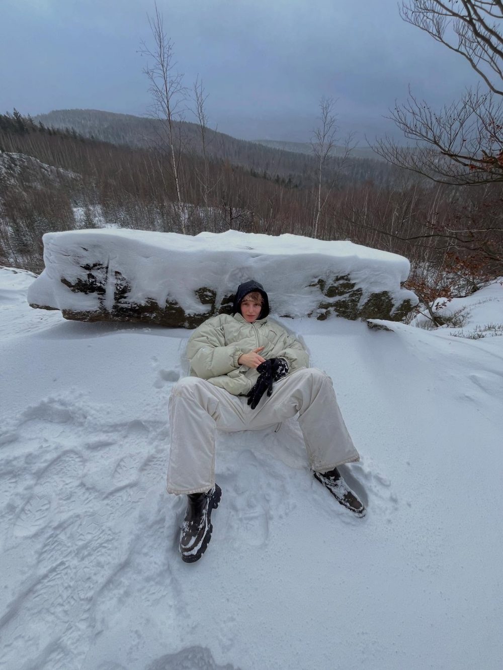 Person in a light jacket and white pants sits on a snowbank with a large snow-covered rock behind, in a wintry forest landscape of bare trees and distant hills under a cloudy sky