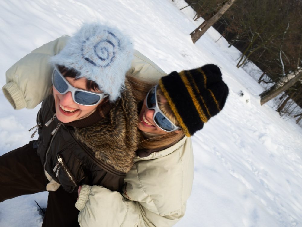 Two women in winter coats and sunglasses laughing and hugging in a snowy park with trees in the background.