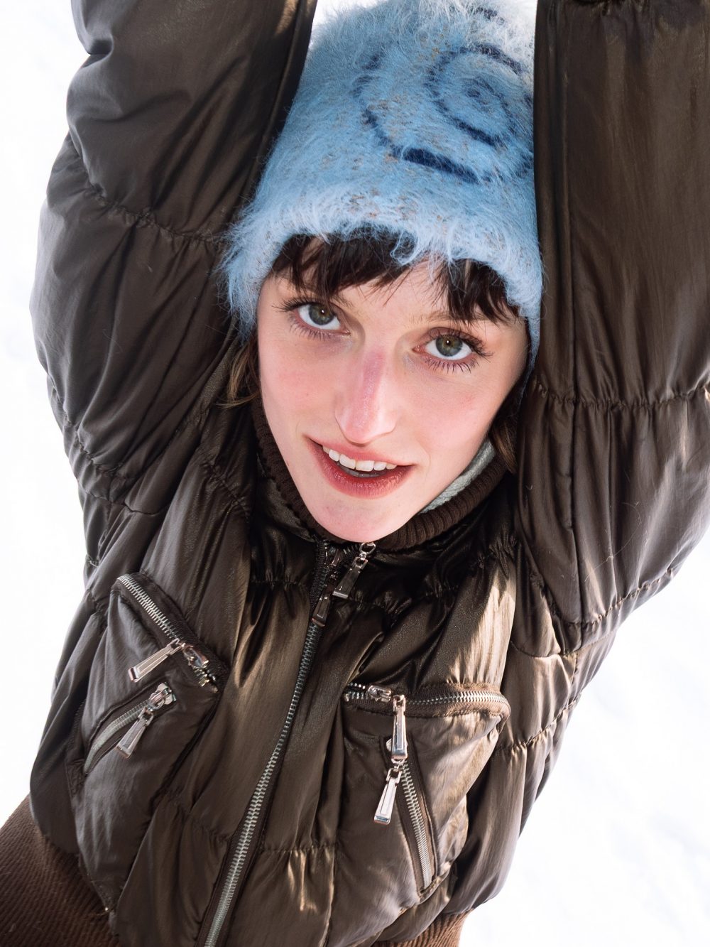 Woman in a brown puffer jacket and light blue fluffy knit hat, smiling at the camera.