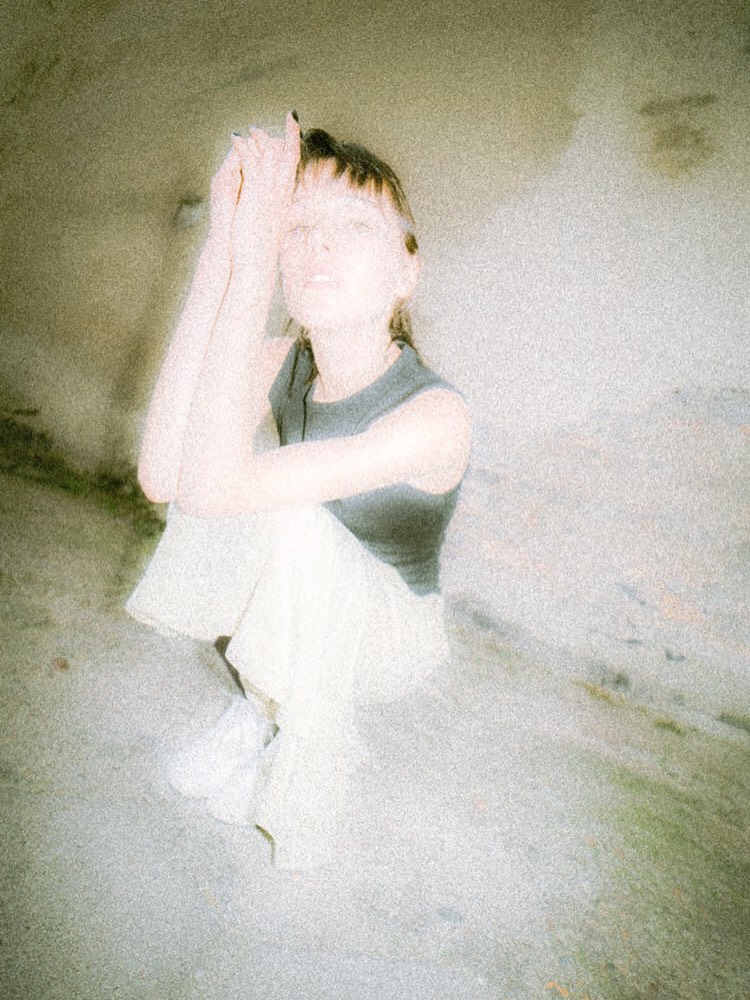 Overexposed, grainy photo of a person crouching with knees up, hands pressed together near the face in a bright, pale setting.