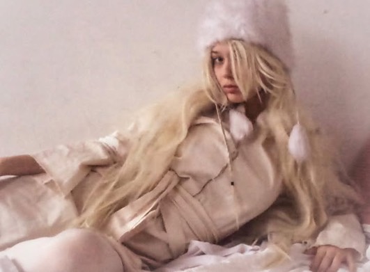Woman with long blonde hair in a cream coat and white fuzzy hat reclining on a bed, looking at the camera.