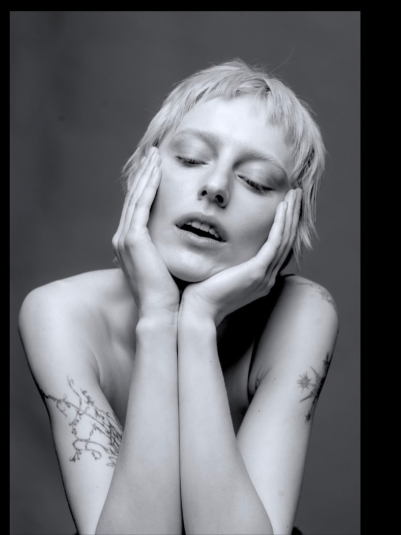 Woman with short blond hair rests her face in her hands, eyes closed, in a monochrome portrait with visible arm tattoos to the elbows.